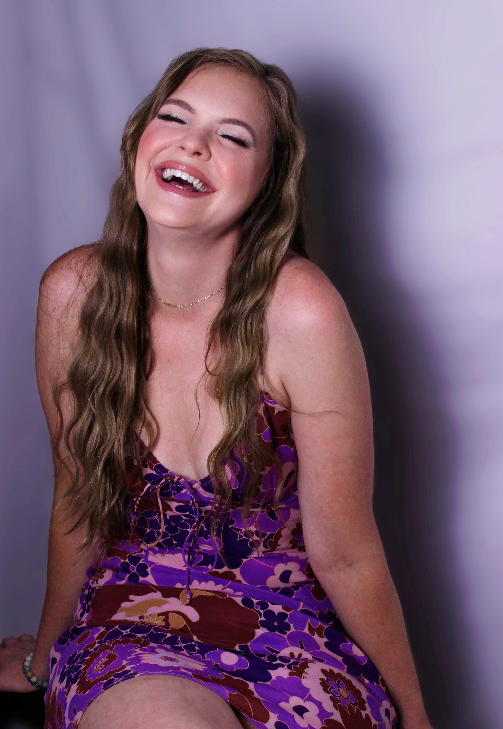Laughing portrait in a purple floral dress against a lilac backdrop with natural shadows