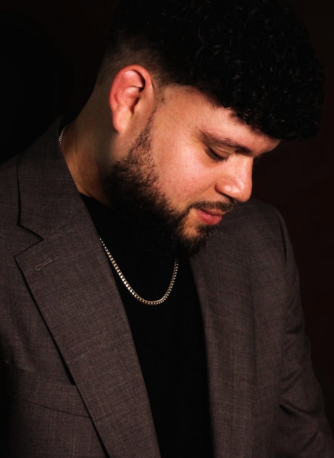 Downward-looking portrait in a dark blazer with a gold chain