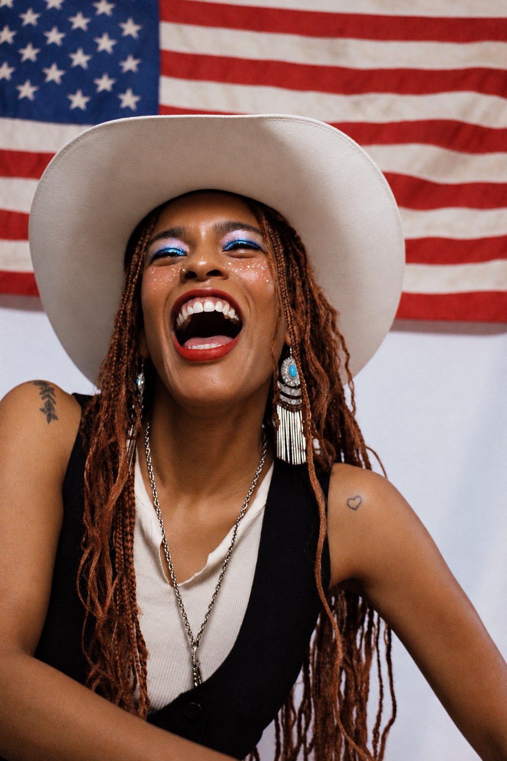 Laughing portrait in a white cowboy hat with colorful eye makeup and an American flag behind