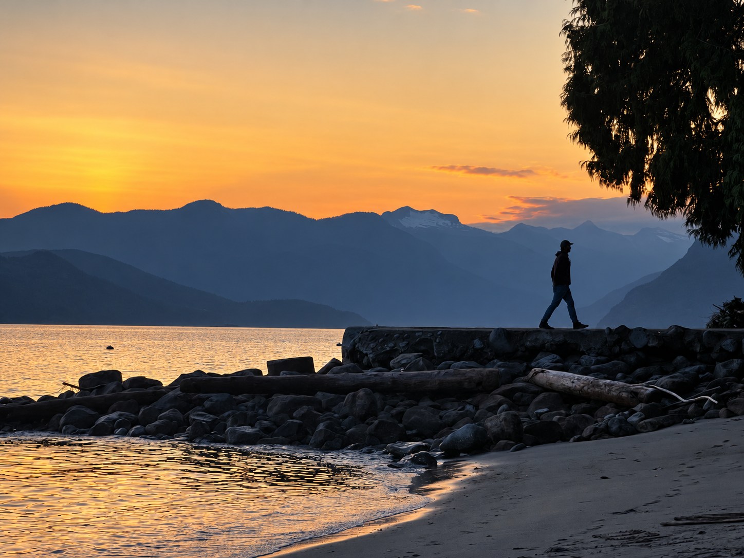 A person walking along a rocky lakeshore beneath layered mountains at dusk