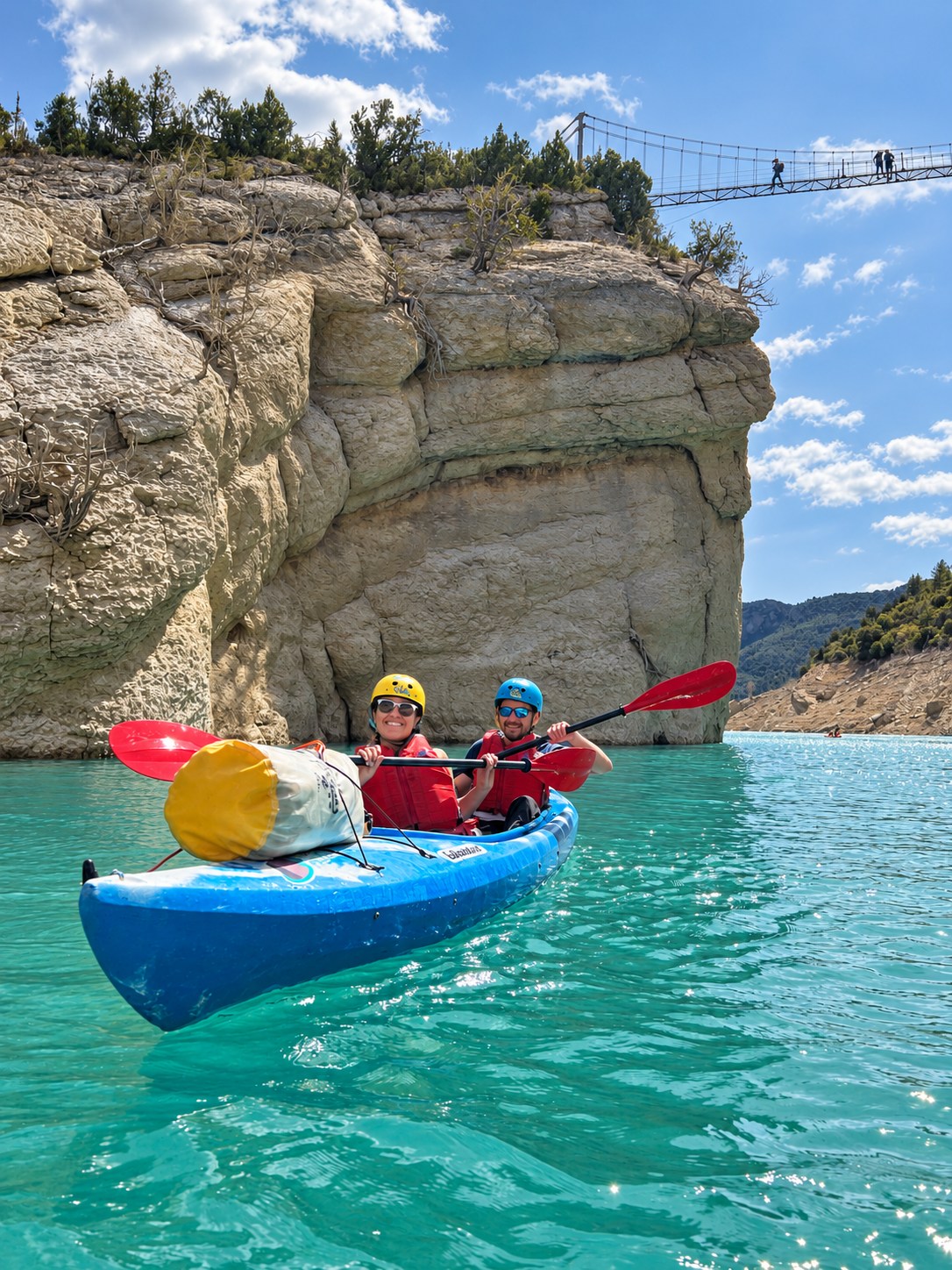 Two kayakers on turquoise water beneath a rocky cliff and suspension bridge